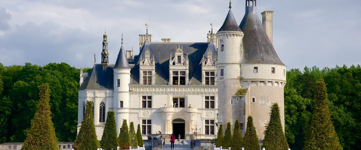A grand castle with multiple towers and turrets, surrounded by a white fence and tall trees.