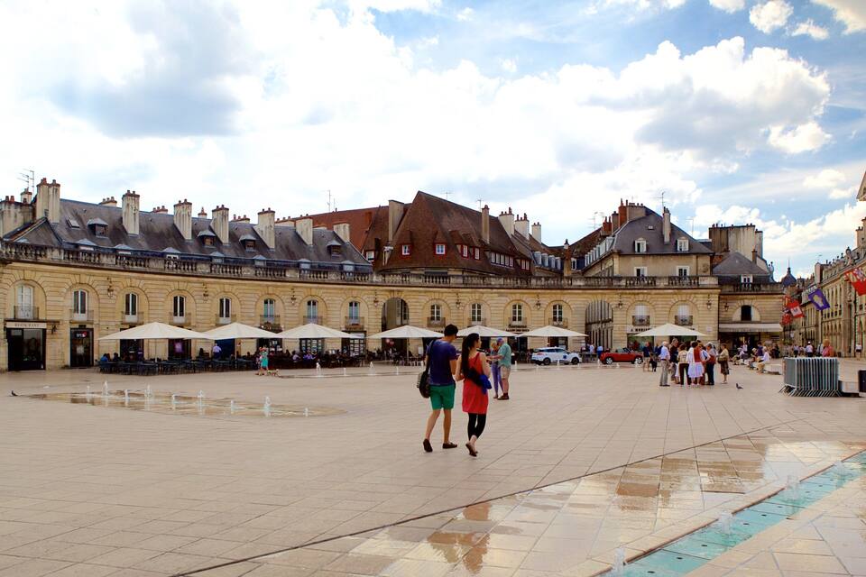 A historic square with a fountain, cafes, and a large building with classical architecture.