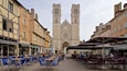 A historic cathedral with twin towers and a large rose window. In front, there are outdoor cafes with tables and chairs.