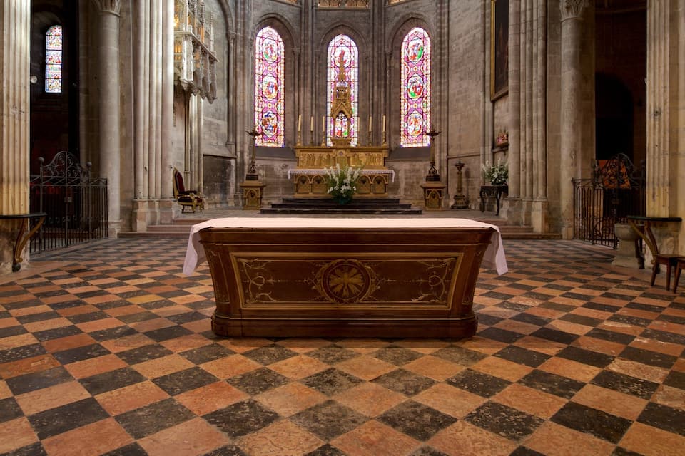 A church interior with a large wooden altar, checkered floor, stained glass windows, and Gothic architecture.