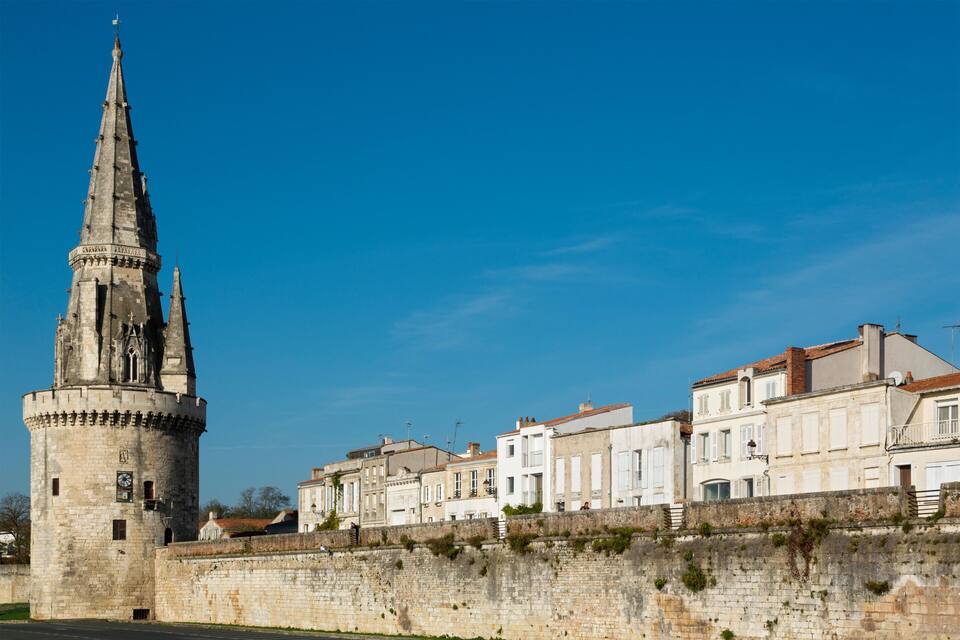A historic tower with a spire, a stone wall, and a row of white buildings with red roofs.