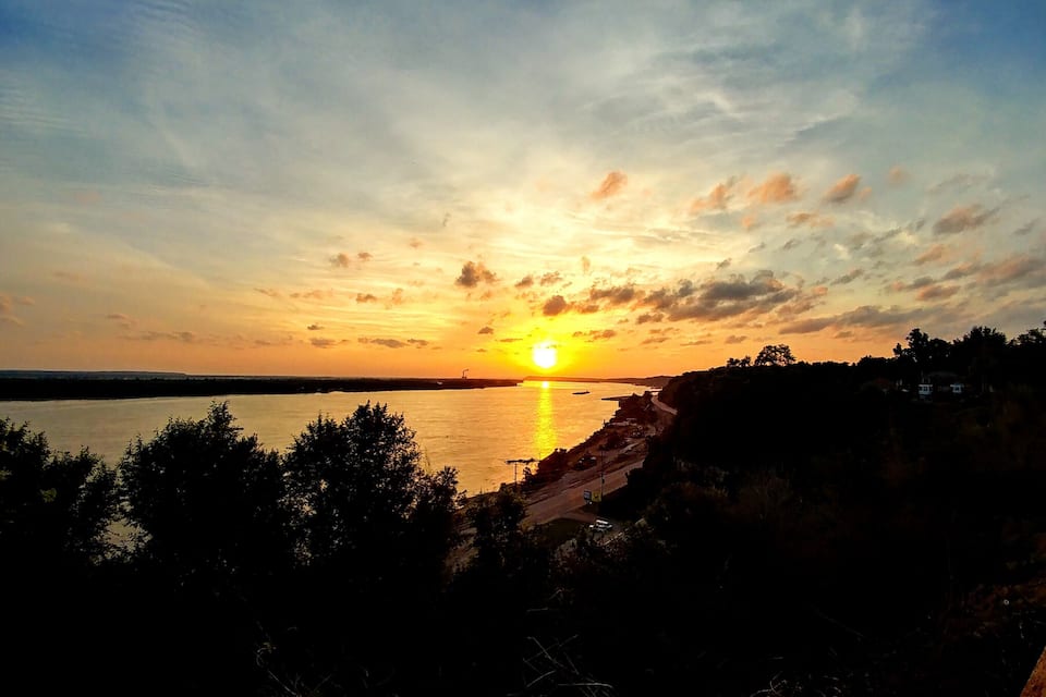 Un atardecer sobre una masa de agua tranquila con costa rocosa y árboles en primer plano.