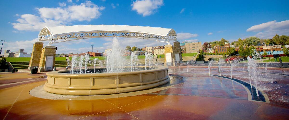 A fountain with water jets under a white arch, surrounded by a paved area with a clear blue sky and buildings in the background.