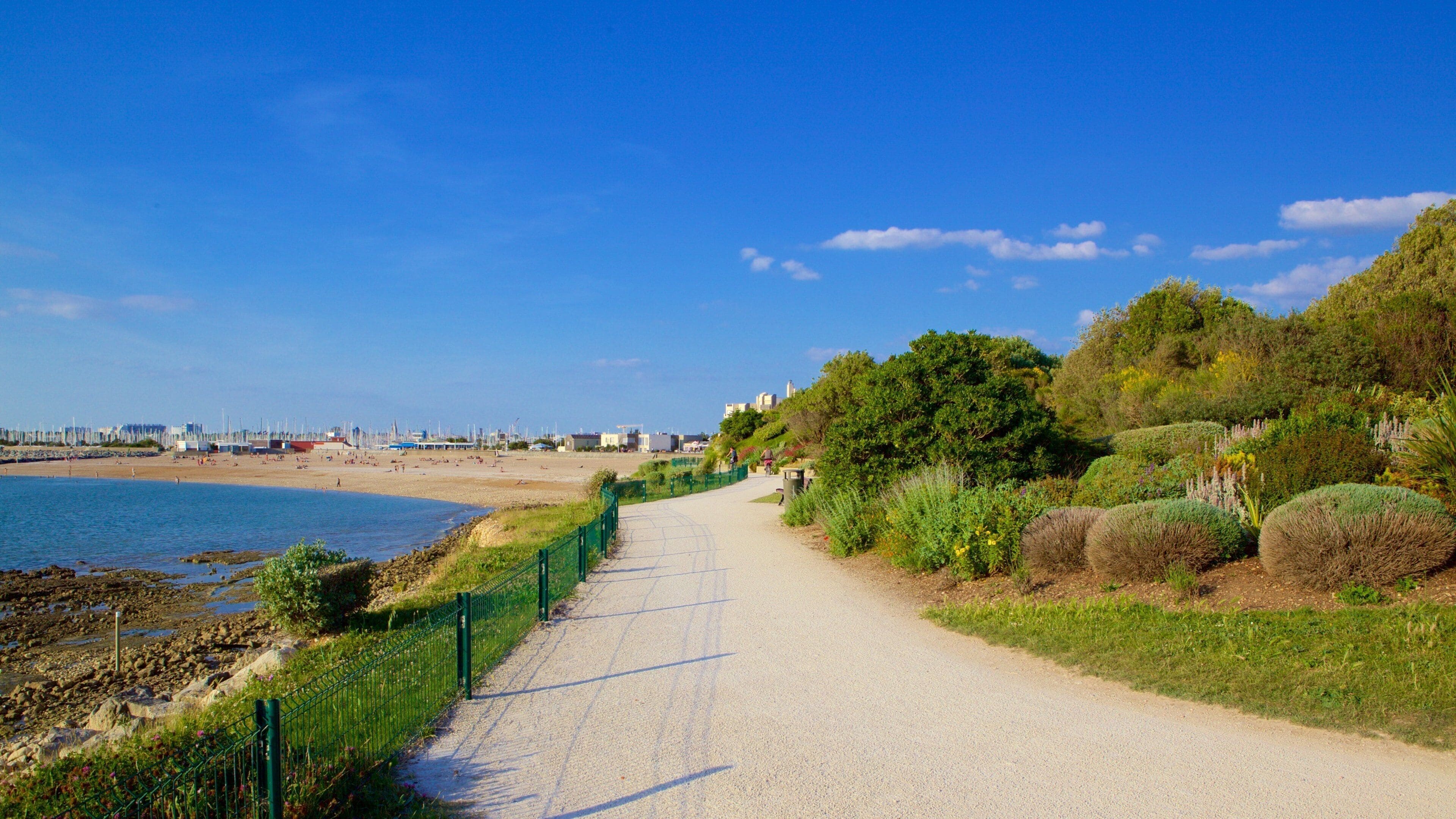 Un sentier côtier bordé d’une clôture verte, avec une plage et une vue sur la ville au loin.