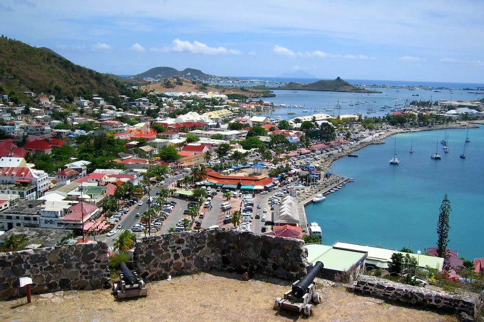 A coastal town with a historic fort, a marina with boats, and a hilly landscape in the background.