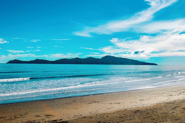 A beach with clear blue water, a sandy shore, and distant mountains under a blue sky.