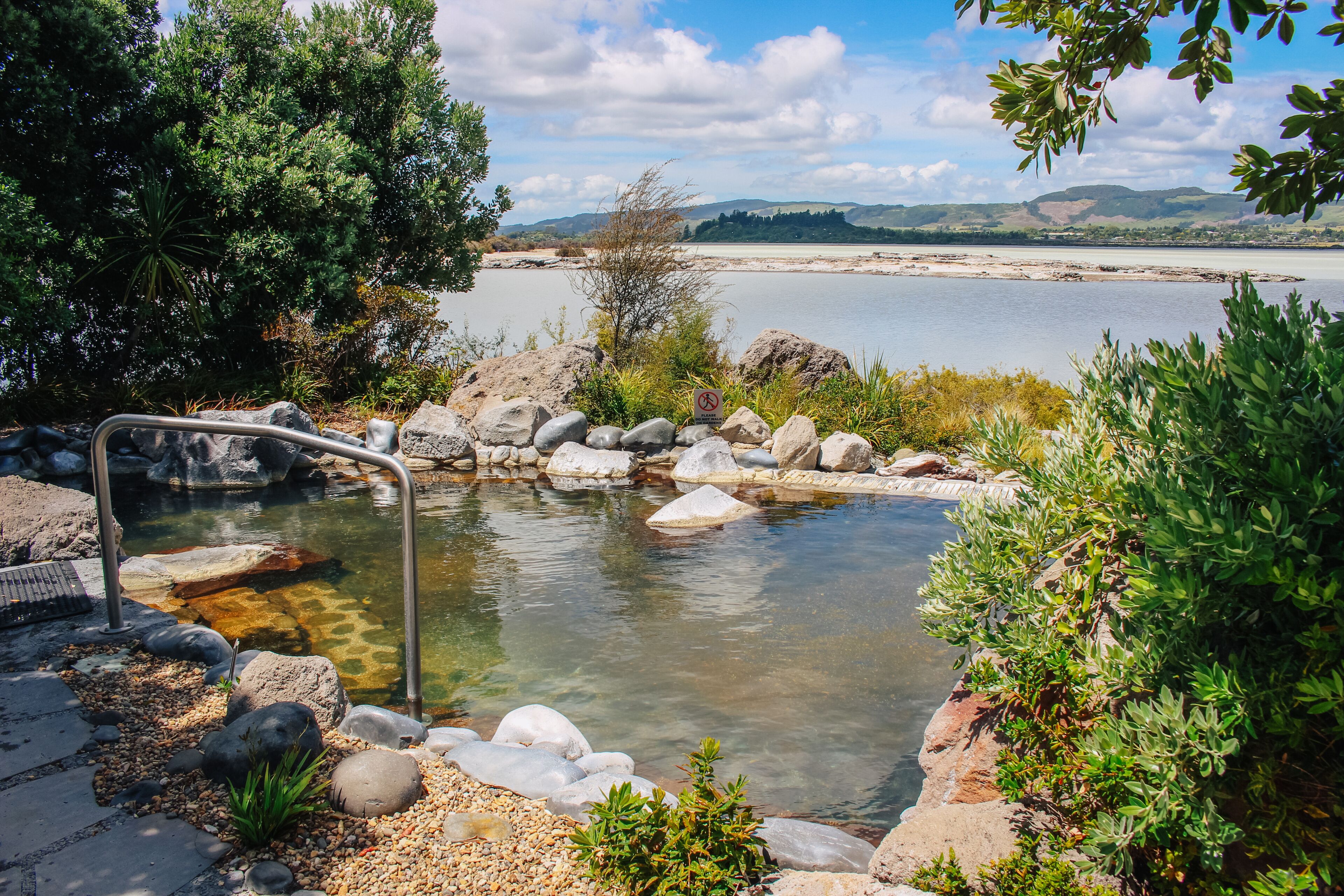 Un tranquilo manantial termal rodeado de rocas y vegetación, con vistas a colinas distantes y un cuerpo de agua.