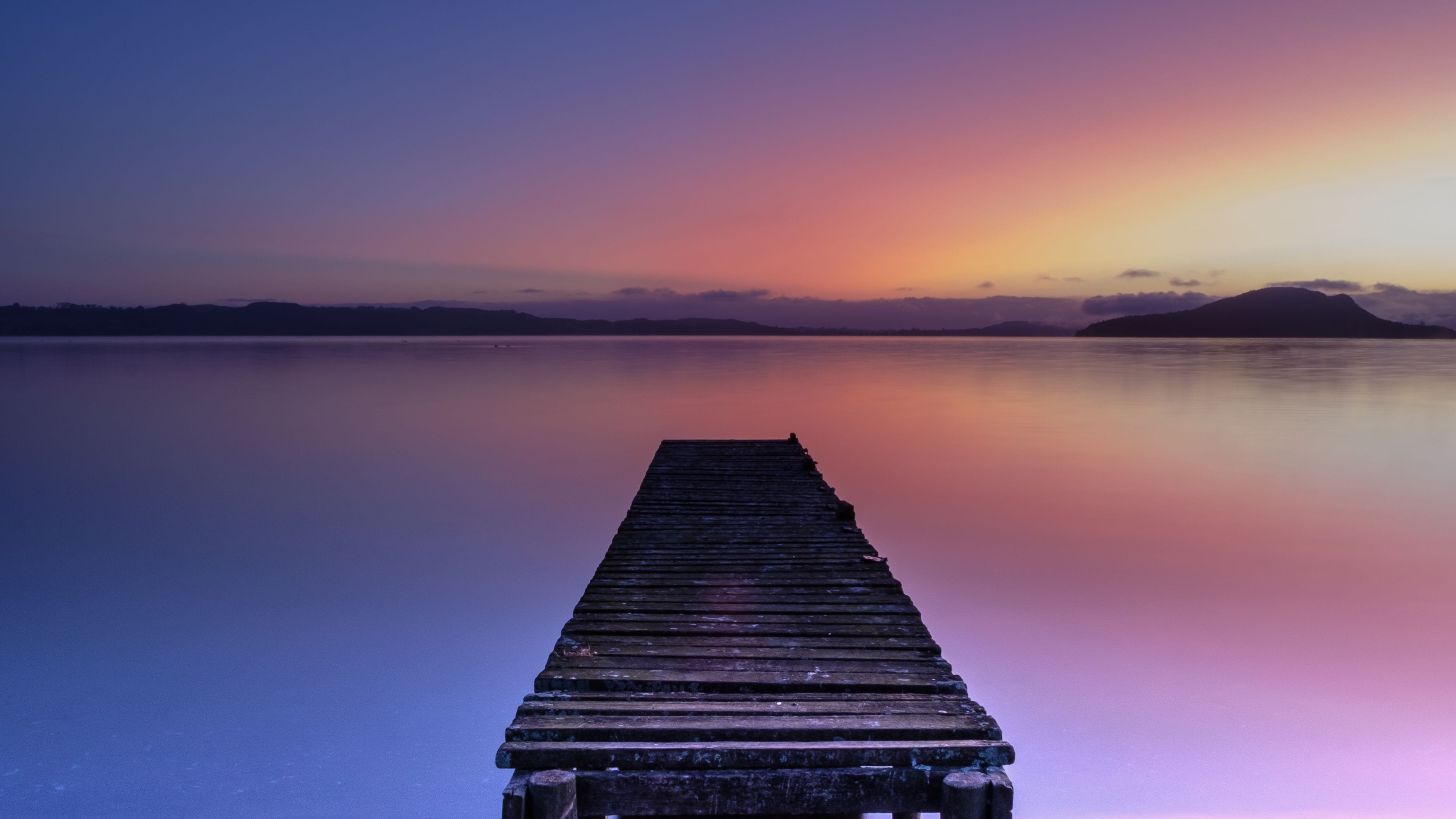 Eine Holzstege in ruhiges Wasser hinein, mit einem Sonnenuntergang im Hintergrund.