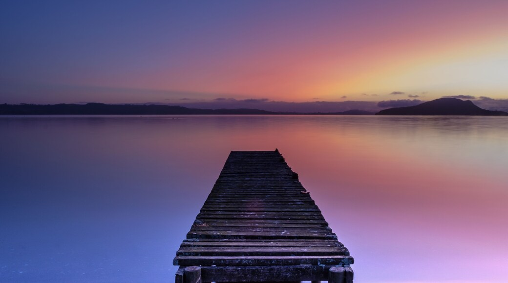 Eine Holzstege in ruhiges Wasser hinein, mit einem Sonnenuntergang im Hintergrund.