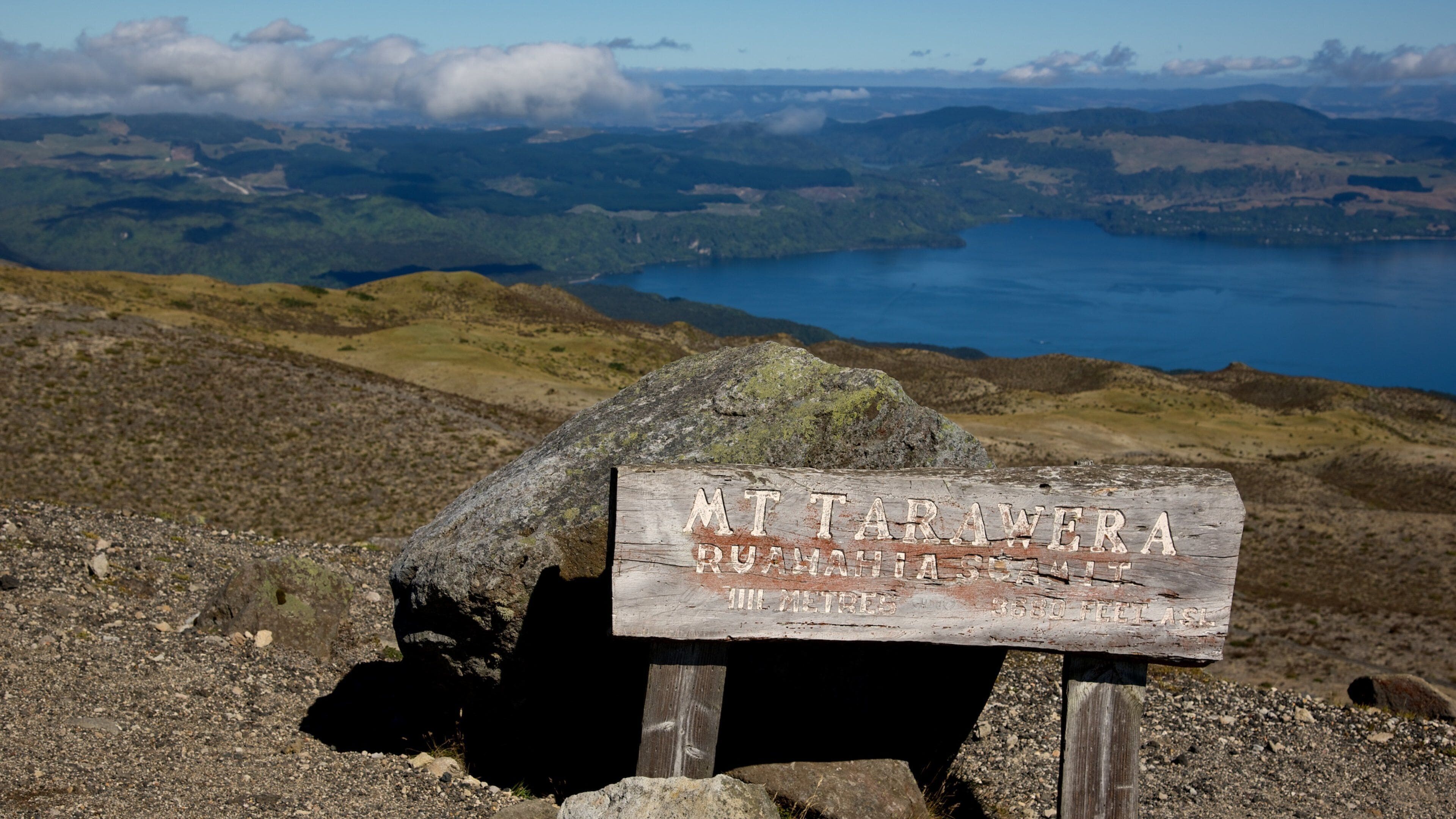 A weathered sign on a rocky outcrop with the text "MT TATARWA RUAHIASCUAMIT III METRES 3580 FEET ASL" and a view of a lake and mountains in the background.