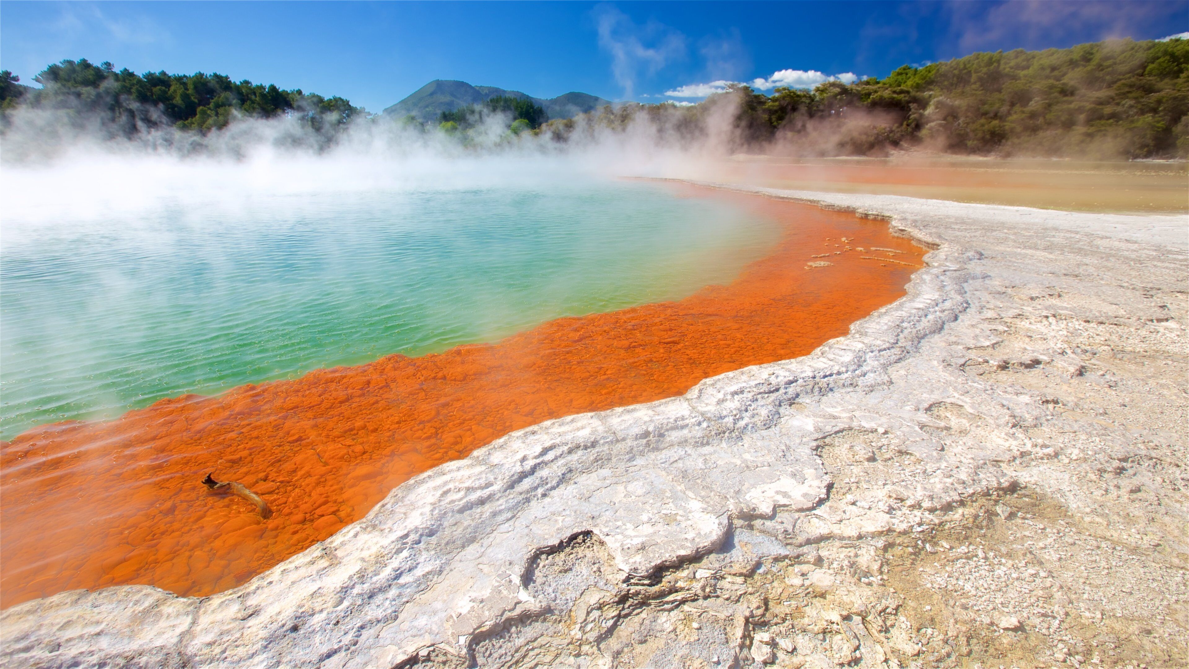 A geothermal hot spring with clear turquoise water, a red mineral deposit, and steam rising from the water.