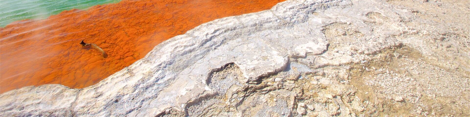 A geothermal hot spring with clear turquoise water, a red mineral deposit, and steam rising from the water.