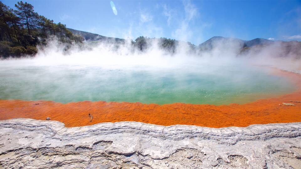 A geothermal hot spring with a red-brown rim and a greenish pool, surrounded by forested hills.
