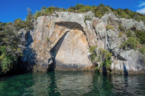 A rock formation with a carved face and a cave entrance surrounded by water and vegetation.
