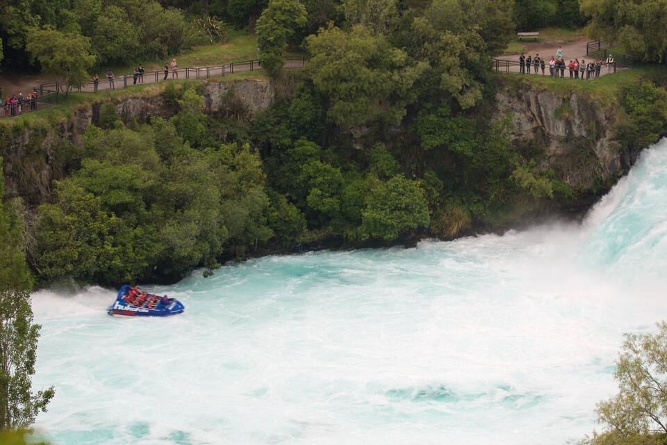 A white water rafting course with a raft in the river, surrounded by dense forest and a viewing platform with people.