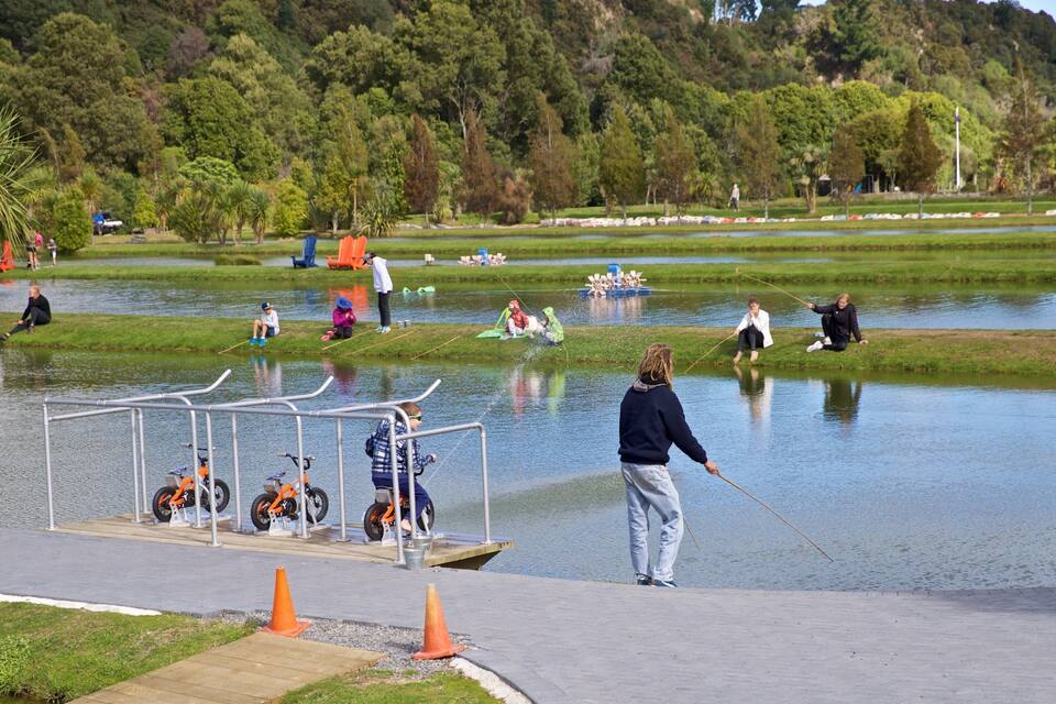 A lakeside park with people fishing and children playing near the water.