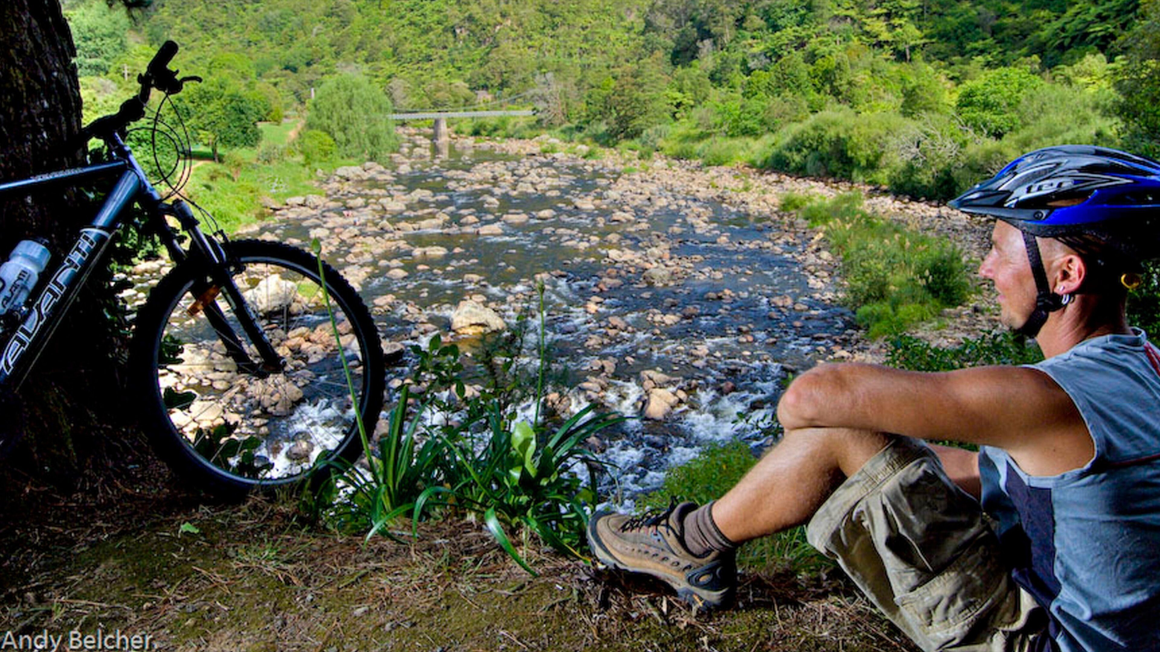 A man sitting by a river with a bicycle leaning against a tree.