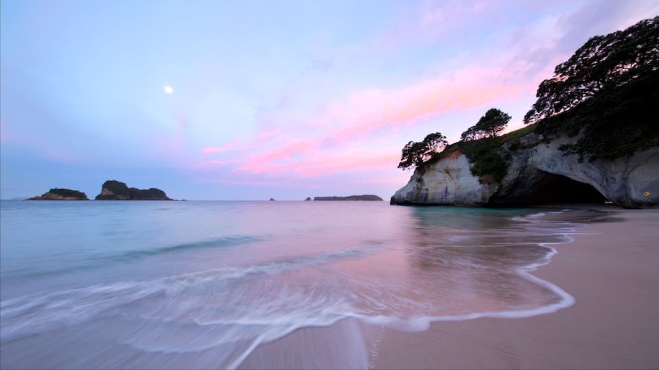 A beach with calm waves, a pink and blue sky, and rocky cliffs.