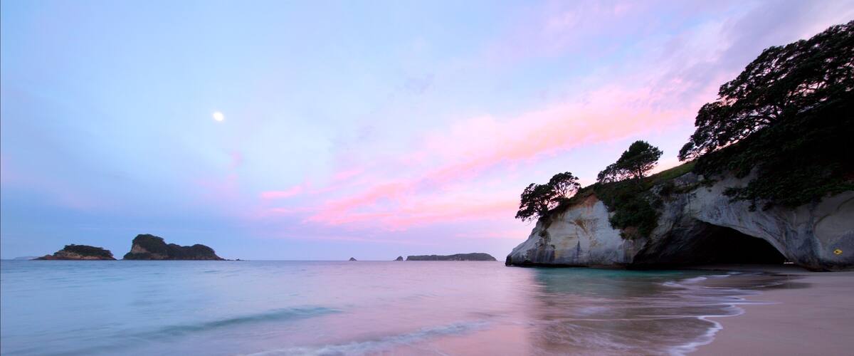 A beach with calm waves, a pink and blue sky, and rocky cliffs.