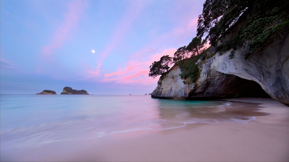 A beach with a large rock formation and a small island in the distance.