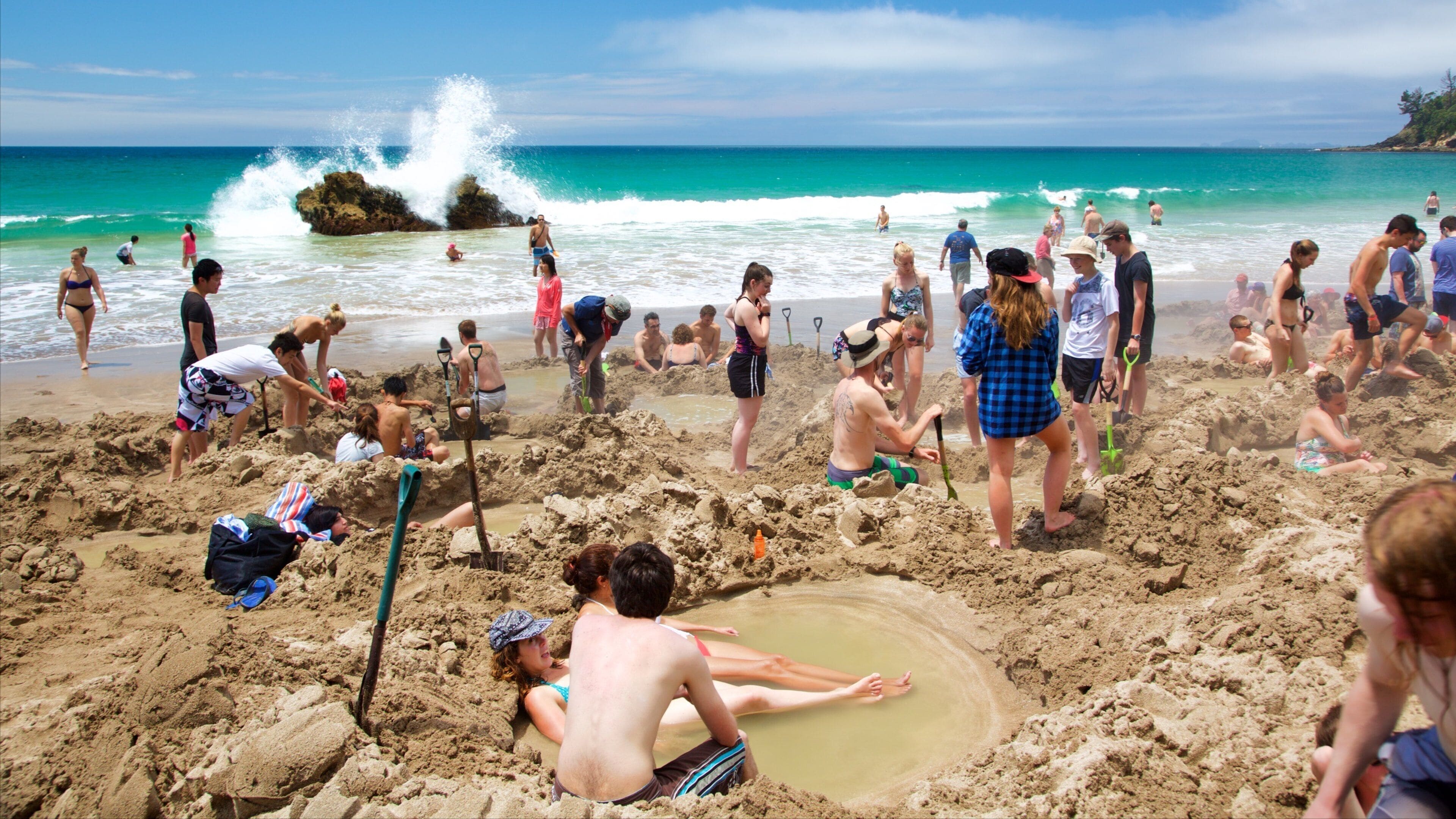 A crowded beach with people playing in the sand and water, and waves crashing against rocks.