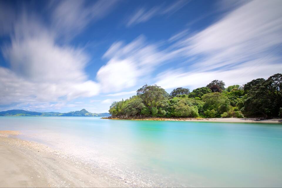 A beach with clear turquoise water, a sandy shore, and a forested hill in the background.