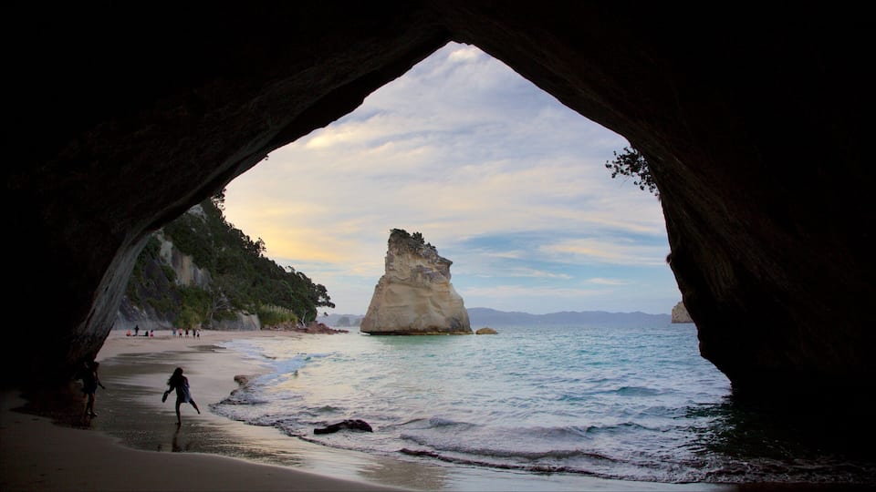 A beach with a large rock formation in the water, viewed through a cave entrance.