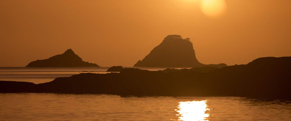 A sunset over a coastal landscape with prominent rock formations silhouetted against the sky.