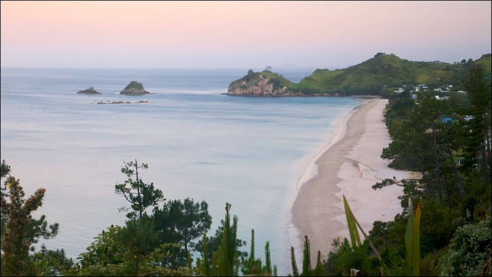 A coastal landscape with a sandy beach, green hills, and rocky outcrops in the sea.