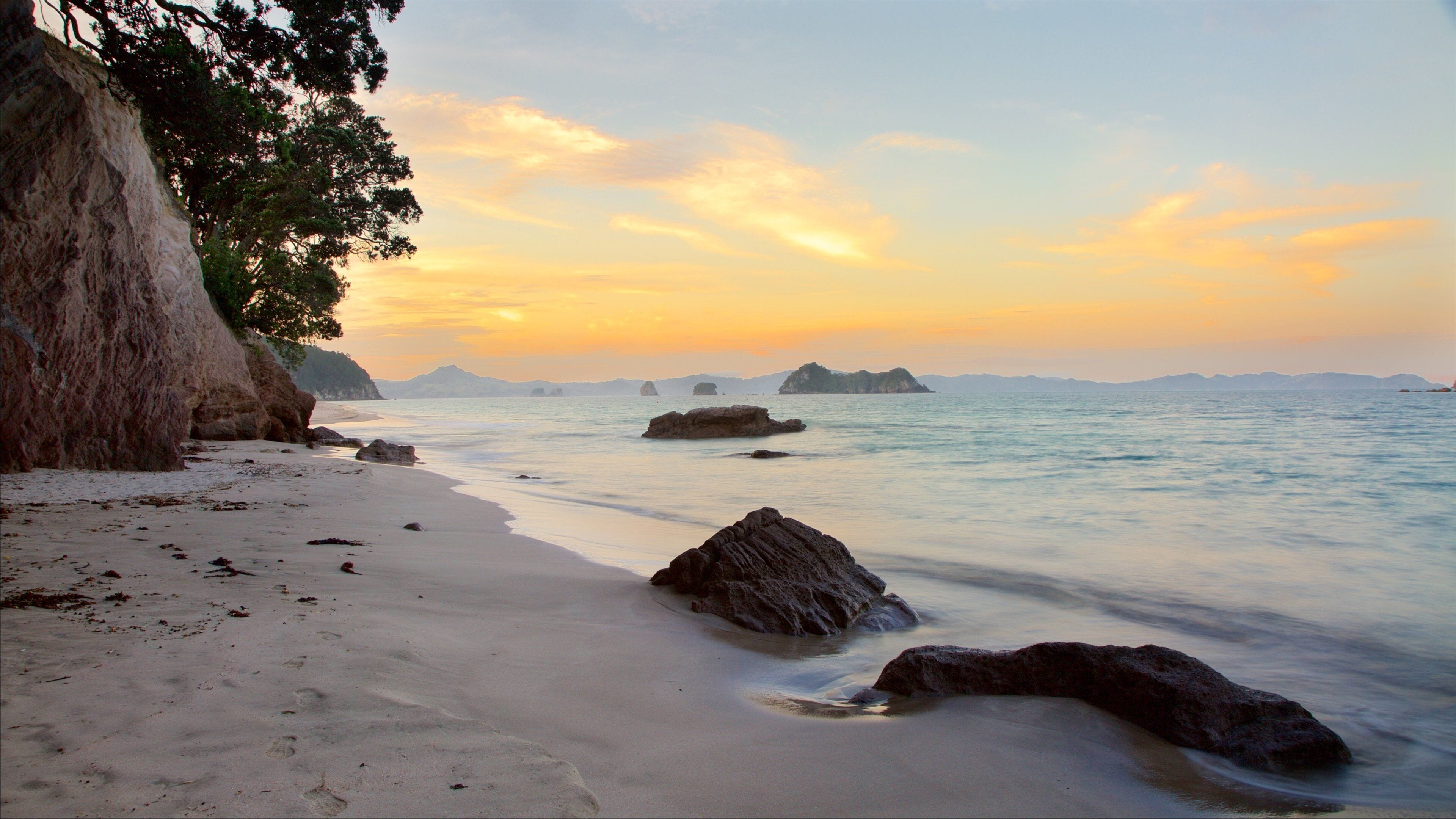 A beach with white sand, large rocks, and distant islands under a sunset sky.