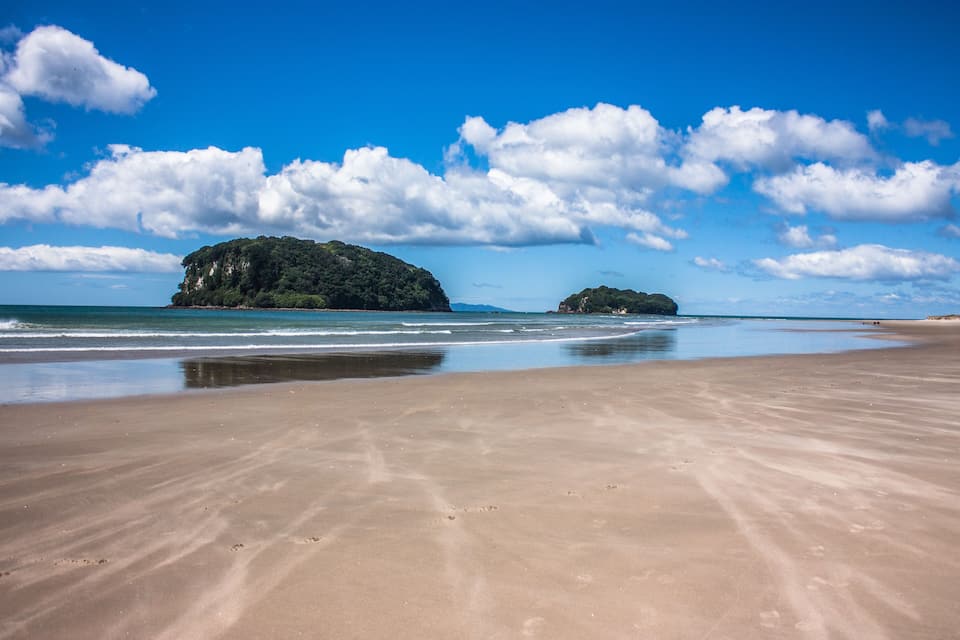 Una playa arenosa con olas tranquilas, un cielo azul despejado y dos islas pequeñas en la distancia.
