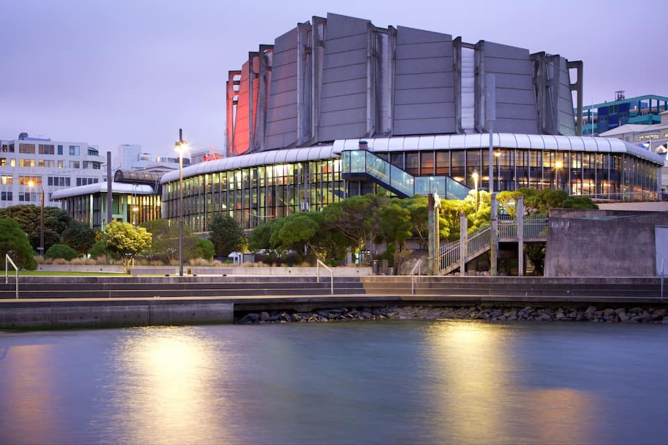 A modern building with a curved glass facade, surrounded by trees and a body of water.