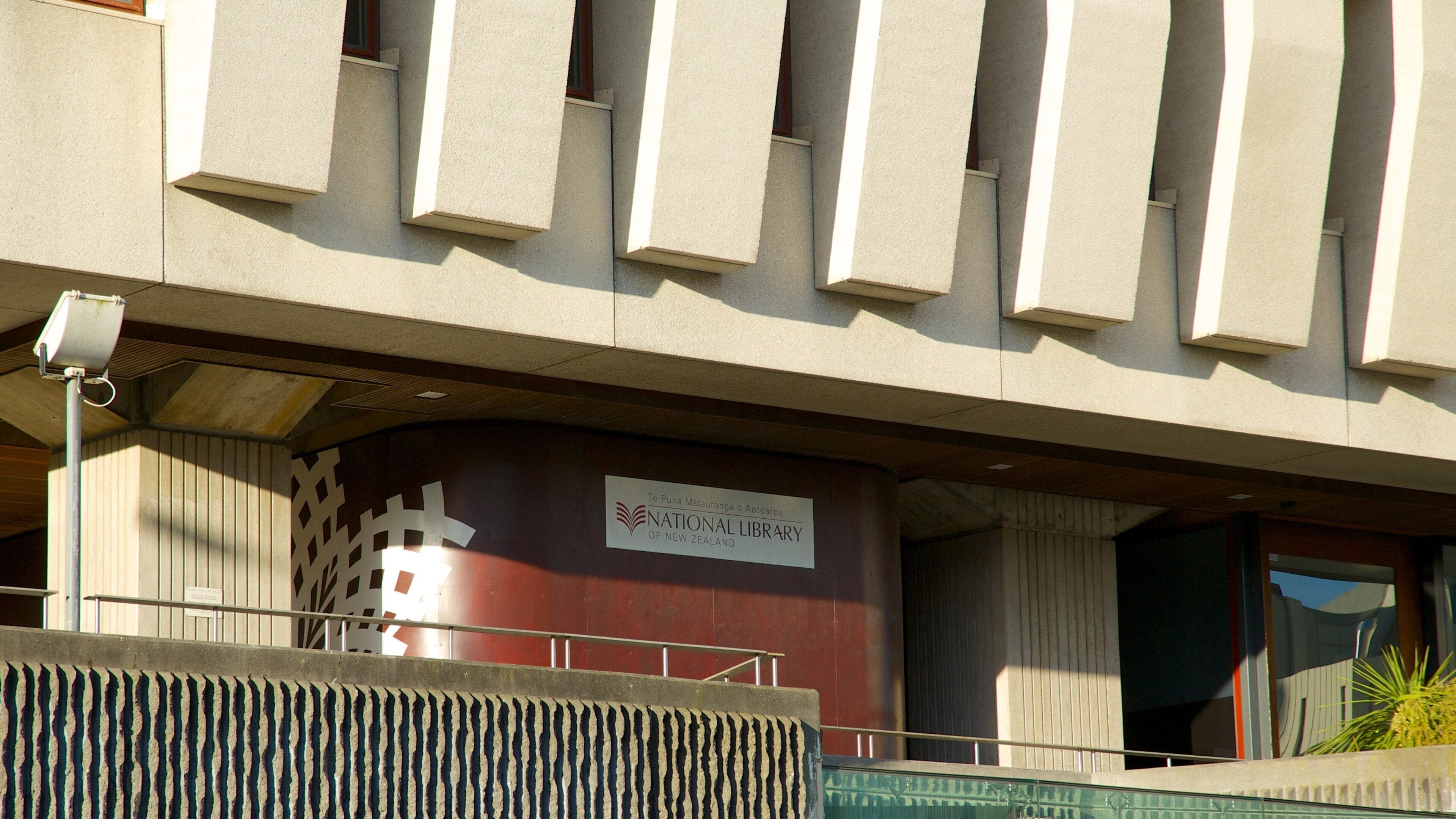 The exterior of the National Library of New Zealand, featuring a modern architectural design with a prominent red and beige facade and a sign indicating its location.