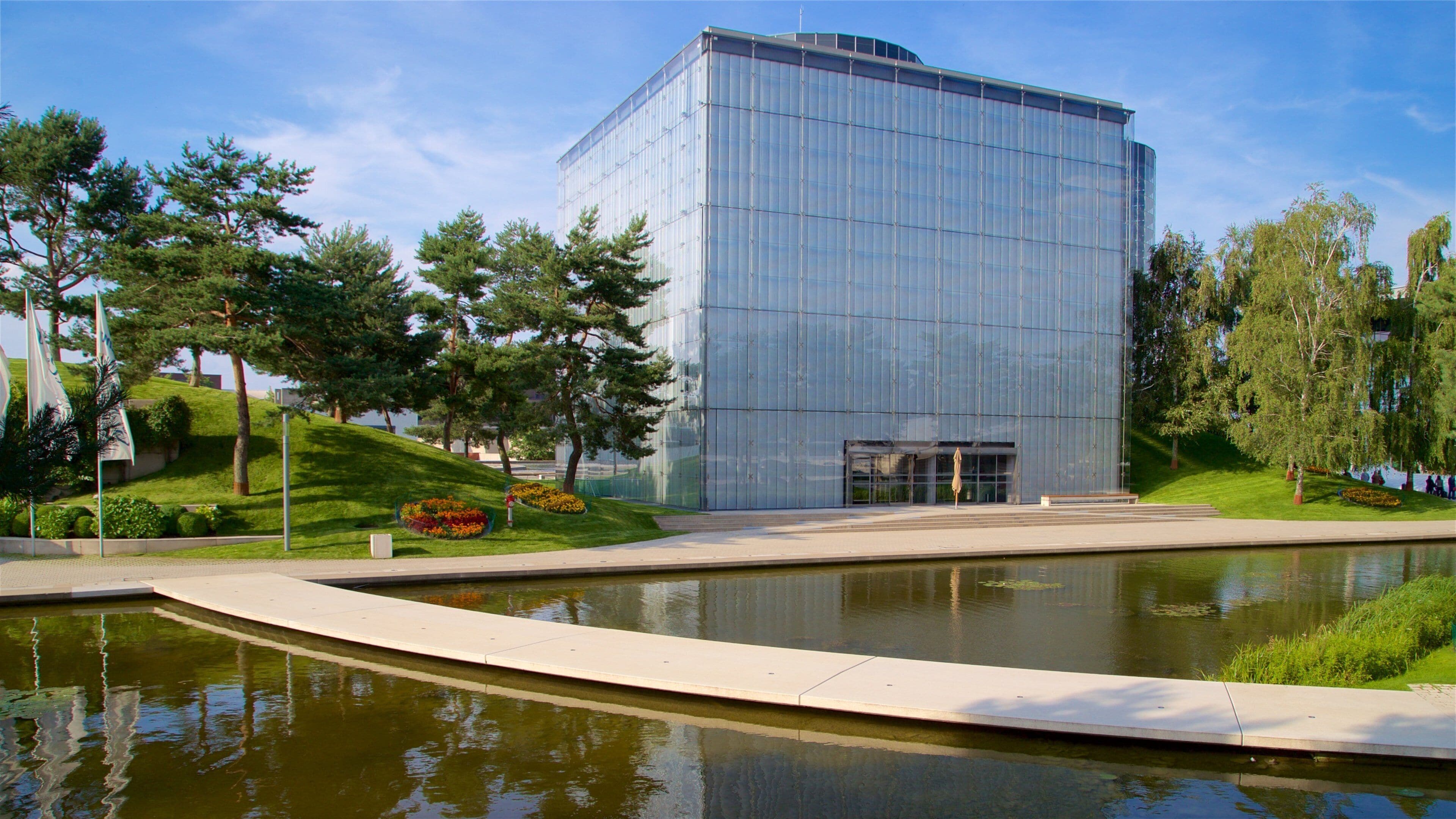 Un bâtiment moderne en verre, doté d’une façade réfléchissante, entouré d’une pelouse bien entretenue, d’un petit étang et de quelques arbres.