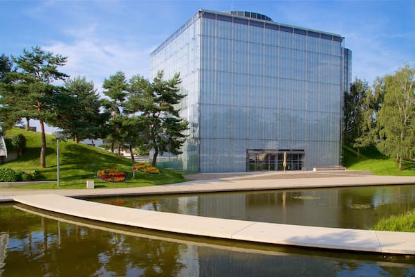 Ein modernes Glasgebäude mit einer spiegelnden Fassade, umgeben von einem gepflegten Rasen, einem kleinen Teich und einigen Bäumen.