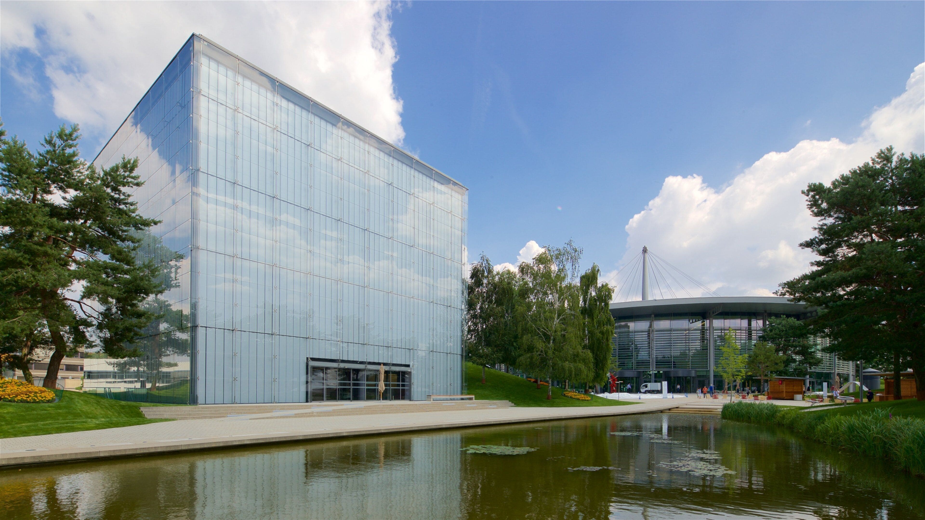 Un edificio moderno in vetro con un riflesso su un corpo d'acqua, circondato da alberi e un cielo sereno.