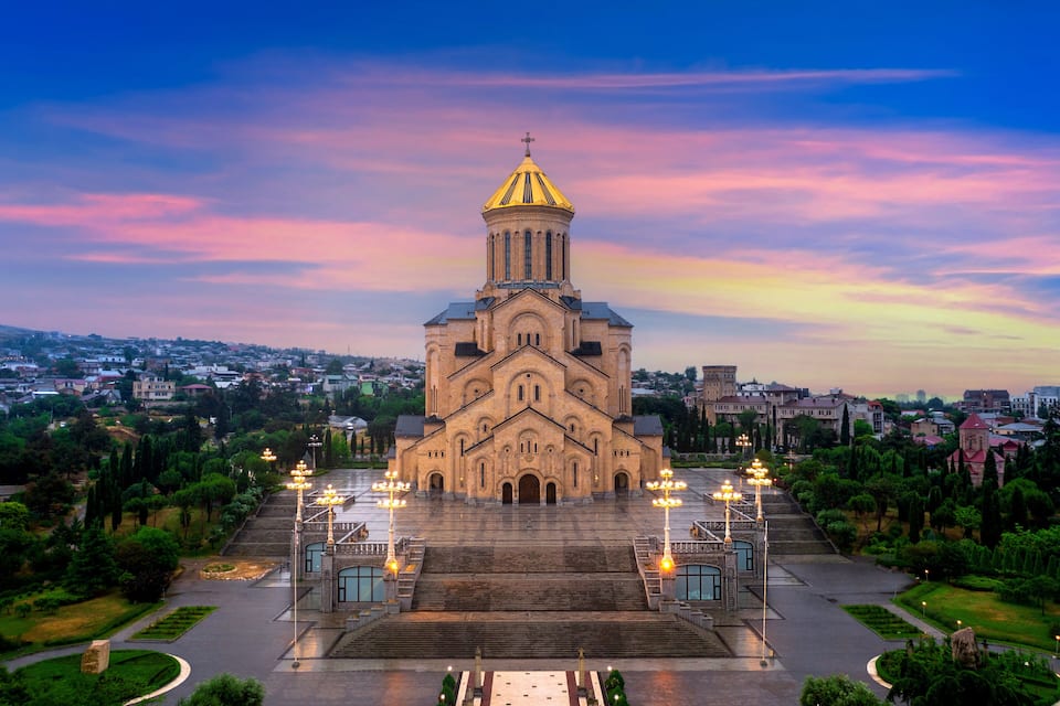 A large church with a golden dome and multiple towers, surrounded by a well-maintained garden and a cityscape in the background.