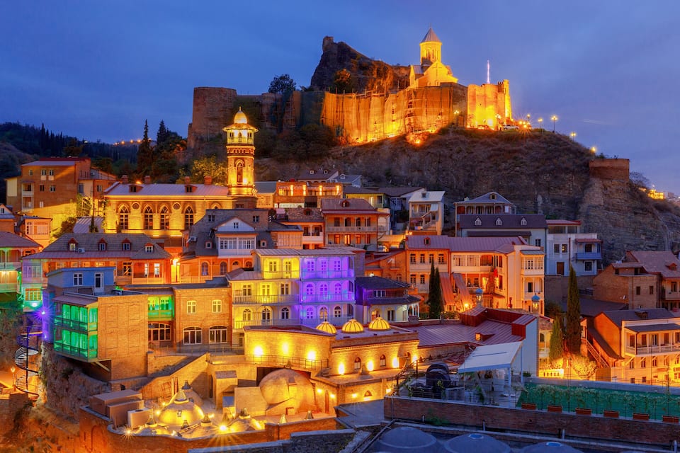 A historic town with illuminated buildings, a prominent castle on a hill, and a domed structure in the foreground.