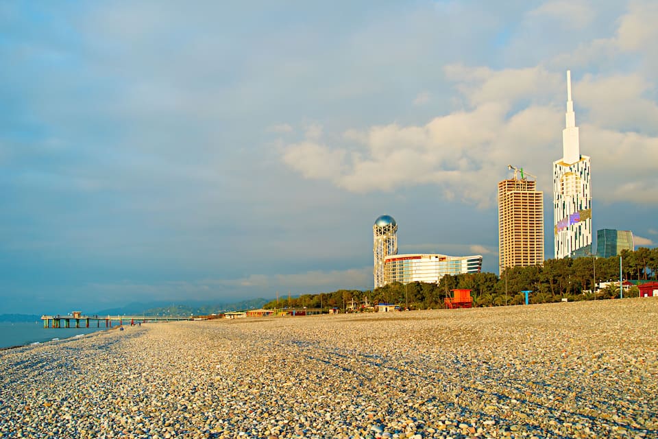 Una playa de guijarros con un muelle que se adentra en el agua, una torre y edificios modernos al fondo.