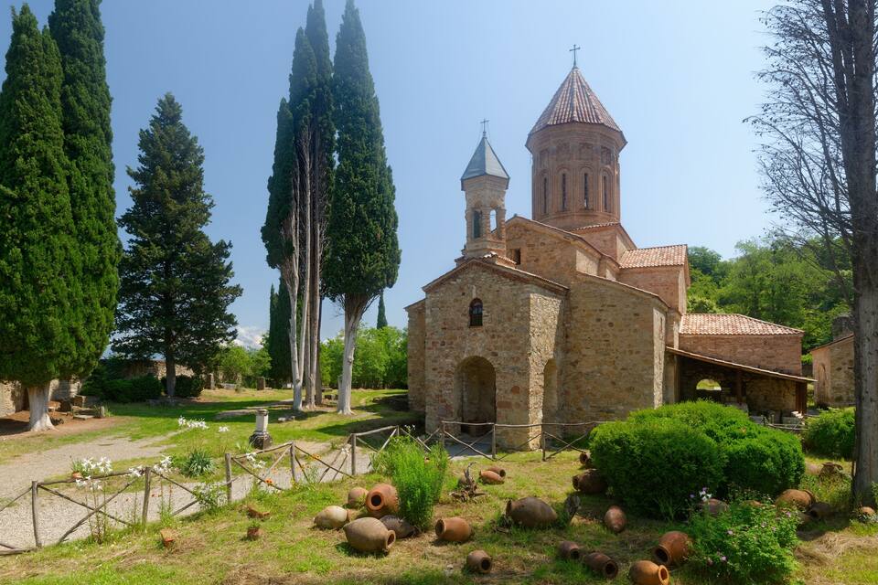 Uma igreja antiga com um campanário, rodeada por ciprestes e uma área gramada com cerâmica.