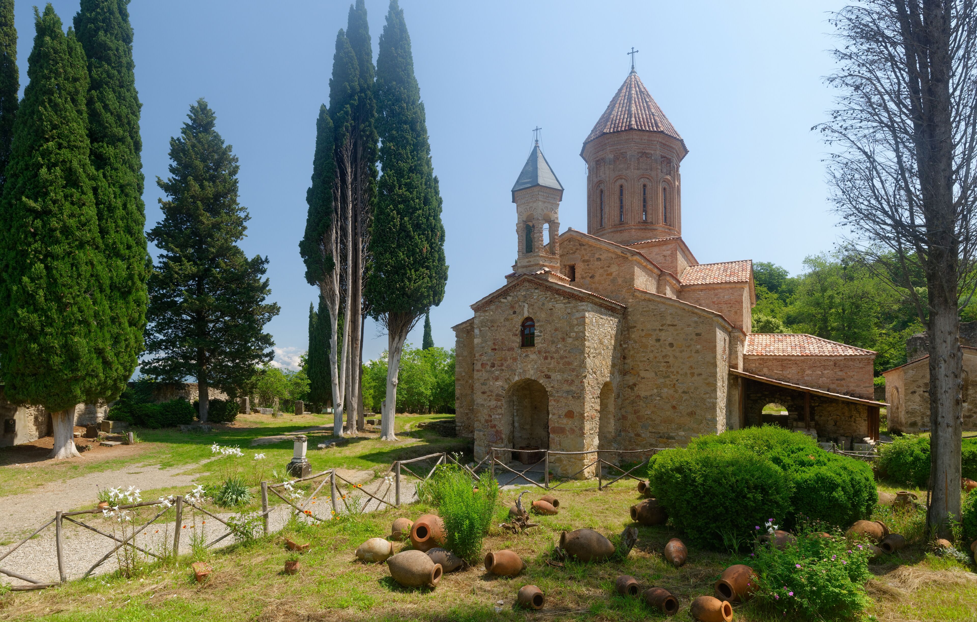 An ancient church with a bell tower, surrounded by cypress trees and a grassy area with pottery.