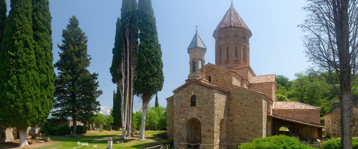 An ancient church with a bell tower, surrounded by cypress trees and a grassy area with pottery.