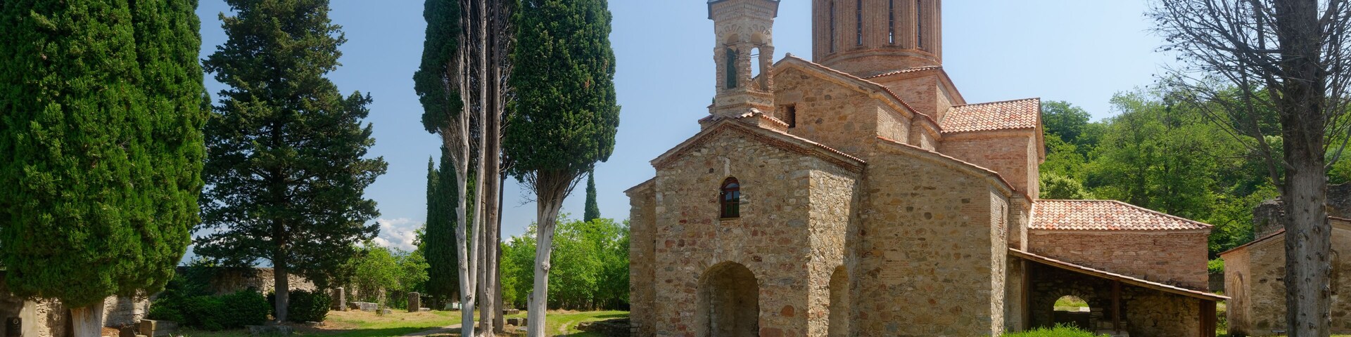 An ancient church with a bell tower, surrounded by cypress trees and a grassy area with pottery.