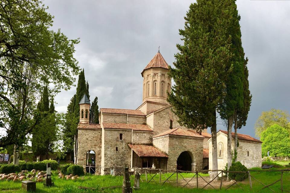 Una iglesia de piedra histórica con un campanario, rodeada de exuberante vegetación y árboles antiguos.
