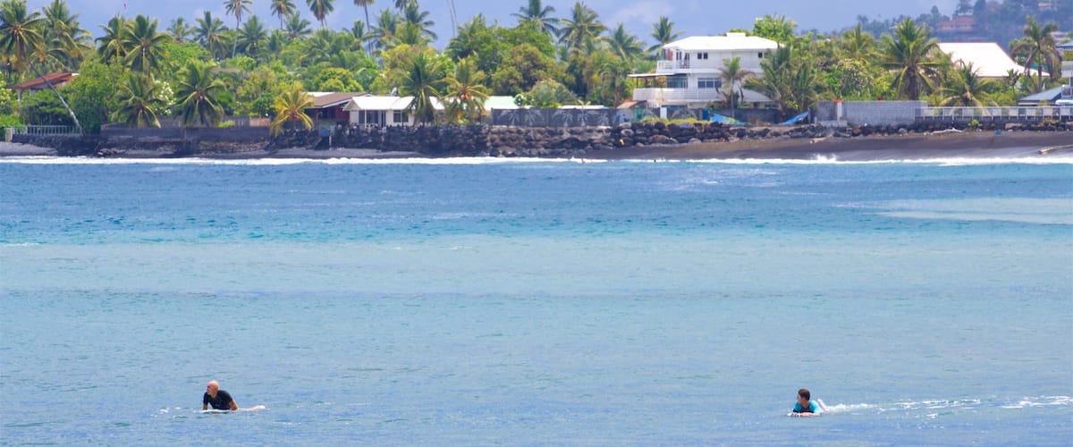 A beach with two people in the water, palm trees, and houses along the shore.