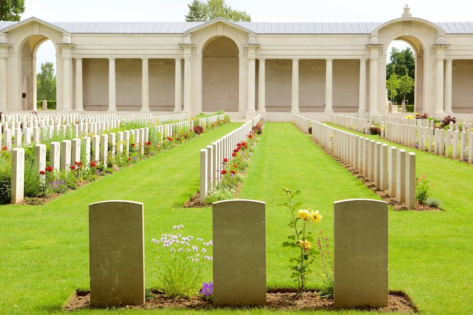 Un cimetière avec des rangées de pierres tombales blanches et, au fond, un bâtiment orné de colonnes.