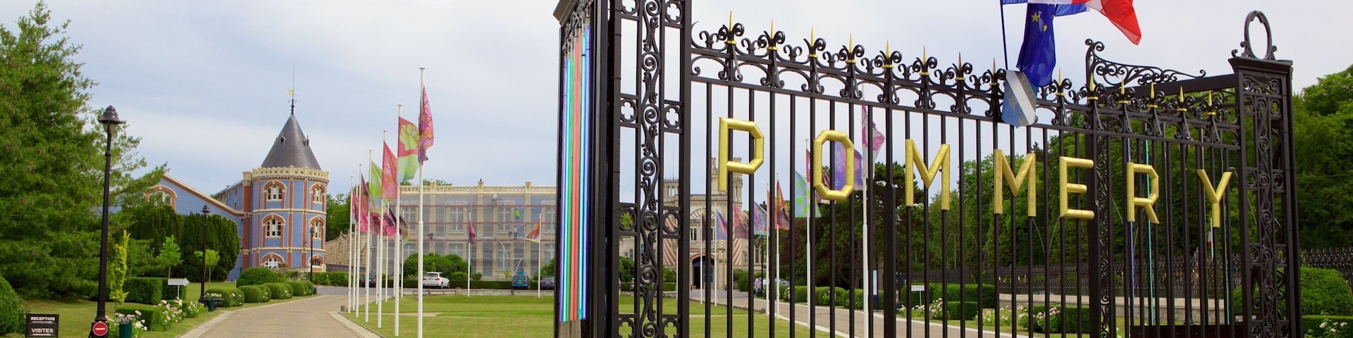 Una reja de hierro forjado con la palabra "POMMERY" en letras doradas, una bandera francesa y un edificio con una torre al fondo.