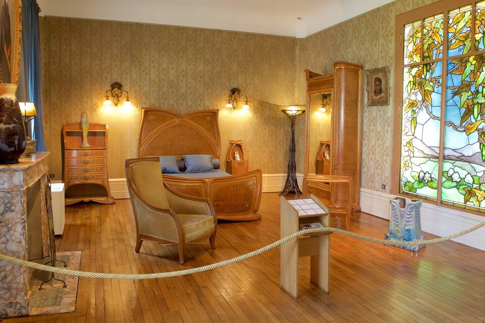 Quarto antigo com cama de madeira, uma cadeira, cômoda e janela de vitral.