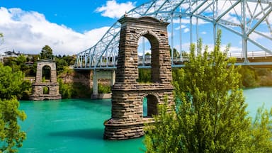 Uma ponte de pedra com estrutura em arco de metal sobre um rio azul-turquesa, cercada por vegetação e casas.