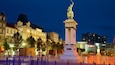 Una plaza urbana con una estatua destacada, edificios históricos y arquitectura moderna.